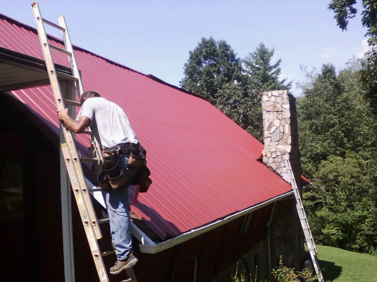Roofer on a ladder beside a red metal roof and stone chimney — Soffit & Fascia Repair by Roofing Brothers in Indiahoma
